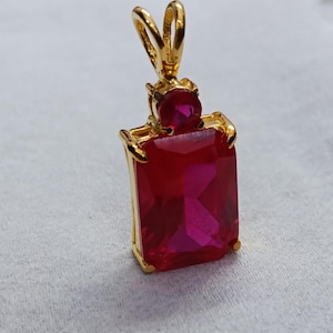 May include: A gold pendant with a large, rectangular, faceted red gemstone. A smaller, round red gemstone is set above the larger stone. The pendant has a gold bail for a chain.