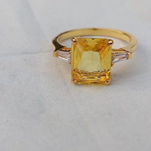 May include: A gold ring with a large, rectangular yellow gemstone. Two smaller, rectangular clear gemstones are set either side of the main stone. The ring has a simple, elegant design.