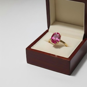 May include: A gold ring with a large, oval-cut pink gemstone, presented in a dark wooden jewelry box with a cream-colored interior. The ring's band is a bright gold color, and the gemstone is a vibrant pink.