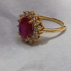 May include: A gold ring featuring a large, oval-cut, ruby-red gemstone surrounded by smaller, clear gemstones. The ring's band is a simple, polished gold. The setting of the central stone has a sunburst design.