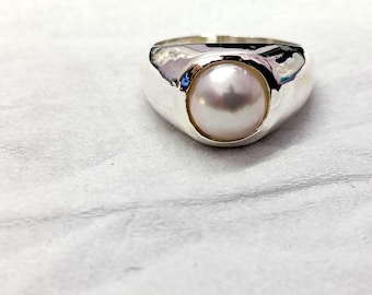 May include: A polished silver ring featuring a single, round, lustrous white pearl set securely in the centre. The band has a smooth, reflective metallic finish that curves around the pearl, creating a clean and simple design for this jewellery piece.