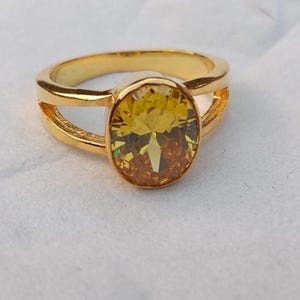 Op de afbeelding: Een goudkleurige ring met een grote, ovaalvormige gele en oranje edelsteen. De ring heeft een gespleten bandontwerp, met twee parallelle banden aan weerszijden van de edelsteen. De ring is geplaatst tegen een witte achtergrond.