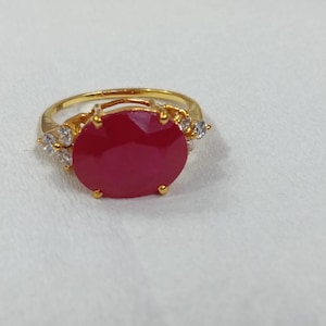 May include: A gold-colored ring featuring a large, oval-cut, deep red gemstone as the centerpiece. The ring is accented with smaller, clear gemstones on either side of the main stone, set against a white background.