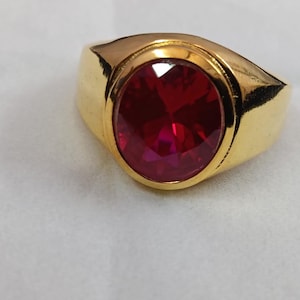 Op de afbeelding: Een goudkleurige ring met een grote, ovaal geslepen, robijnrode edelsteen. De ring heeft een gladde, gepolijste band en een bezel-zetting die de levendige steen stevig vasthoudt. De ring staat tegen een witte achtergrond.