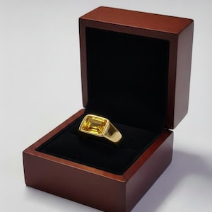 May include: A gold ring with a rectangular yellow gemstone, presented in an open, dark wooden jewellery box with a black velvet lining. The box is set against a neutral background.