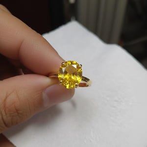 May include: A gold ring featuring a large, oval-cut yellow gemstone. The ring's band is a simple, polished gold. The gemstone is a vibrant yellow color, and the ring is held up against a white background.