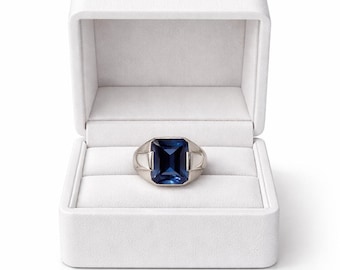 May include: A silver ring with a large, rectangular, deep blue gemstone, presented in an open white jewelry box. The ring has a classic design with a wide band and a prominent stone setting.