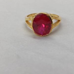 Könnte beinhalten: Ein goldfarbener Ring mit einem großen, ovalen, facettierten roten Edelstein in der Mitte. Die Ringschiene weist auf beiden Seiten ein geteiltes Design auf, das zur zentralen Fassung führt, in der der leuchtende, durchscheinende rote Stein sitzt.