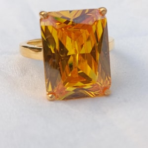 Op de afbeelding: Een goudkleurige ring met een grote, rechthoekige, geel-oranje edelsteen. De edelsteen is het middelpunt, met een facetgeslepen oppervlak dat het licht vangt. De ring heeft een eenvoudige, gepolijste gouden band.