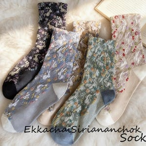 May include: Five pairs of patterned socks in various colors, including black, gray, and beige, with floral and textured designs. The socks are arranged on a fluffy white surface, with an open book in the background. The text "Ekkachai Siriananchok Socks" is visible.