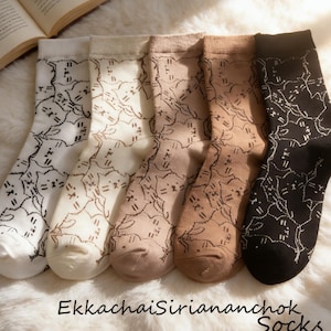 Op de afbeelding: Vijf paar sokken in verschillende neutrale kleuren, elk met een herhaald kattengezichtpatroon. De sokken variëren van gebroken wit tot zwart, met de tekst "EkkachaiSiriananchok Socks" onderaan.