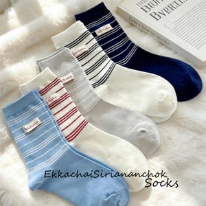 Può includere: Una collezione di calzini a righe in vari colori, tra cui blu, rosso, grigio e blu navy. Ogni calzino presenta una piccola etichetta con testo. I calzini sono disposti su una superficie morbida e bianca. Il testo "EkkachaiSiriananchok Socks" è visibile.