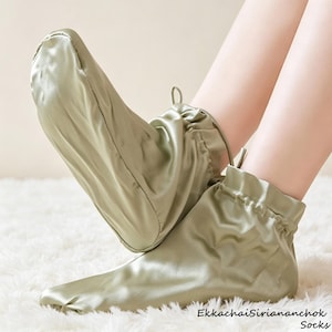 Op de afbeelding: Een paar saliegroene satijnen sokken met een gladde, glanzende afwerking. De sokken hebben een geplooide, elastische boord bij de enkel en een afgeronde teen. De tekst EkkachaiSiriananchok Socks staat rechtsonder in de afbeelding gedrukt.