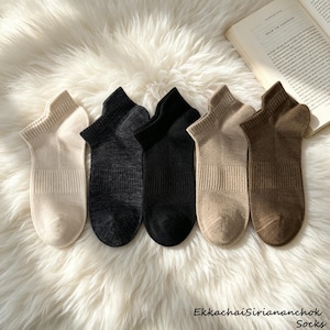 May include: A set of five ankle socks in a row, featuring a ribbed cuff and a solid knit texture. The colors include cream, charcoal gray, solid black, light beige, and dark brown. Each sock has a reinforced heel and toe area with a consistent design.