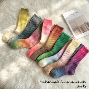 Peut inclure: Une collection de chaussettes côtelées colorées dans diverses teintes, dont le jaune, le gris, le rose, l'orange, le vert et le violet. Les chaussettes sont disposées sur une surface blanche et duveteuse. Le texte "EkkachaiSiriananchok Socks" est visible en bas.