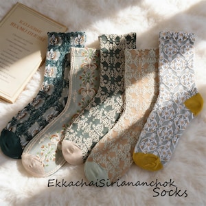 May include: Five pairs of patterned socks in various colors, including teal, sage green, and beige, with intricate lace and floral designs. The socks have different cuff and toe colors, and are displayed on a fluffy white surface. The text "EkkachaiSiriananchok Socks" is visible.