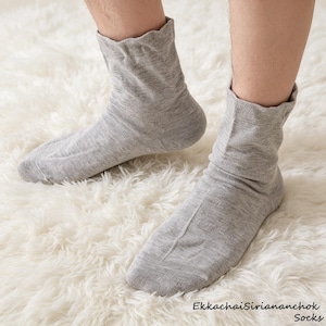 May include: A pair of light grey crew-length socks with a fine knit texture and a ribbed cuff. The socks feature a subtle heathered appearance and are worn on the feet. The text EkkachaiSiriananchok Socks is printed in the bottom right corner.