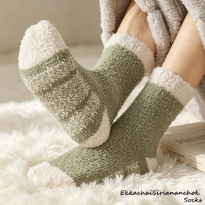 Könnte beinhalten: Ein Paar flauschige Socken mit einem salbeigrünen Körper und cremefarbenen Zehen, Fersen und Bündchen. Die Socken haben eine weiche, strukturierte Strickoptik und werden an den Füßen getragen, wobei der Text EkkachaiSiriananchok Socks sichtbar ist.