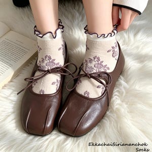 May include: A pair of dark brown leather ballet flats with a square toe and a thin tie detail across the top. The shoes are worn with cream-colored ankle socks featuring a purple floral pattern and a ruffled edge with a dark purple trim.
