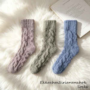 May include: Three cable-knit socks in lavender, grey-green, and blue. The socks are displayed on a fluffy white surface. The text "EkkachaiSiriananchok Socks" is visible at the bottom of the image.