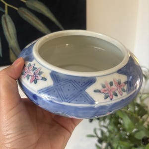 May include: A small, round ceramic bowl with a white interior and a blue and white exterior featuring a geometric cross-hatch pattern and small pink floral motifs. The bowl has a wide, open top and a smooth, glazed finish on the outer surface.