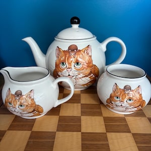 May include: A white ceramic tea set comprising a teapot, creamer jug, and sugar bowl. Each item is adorned with a playful design of an orange tabby cat with a mouse on its head. The set has black trim and a black knob on the teapot lid.