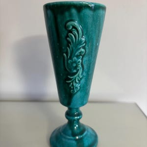 May include: A teal ceramic goblet with a flared top and a pedestal base. The goblet features a raised floral design on the front. The glossy finish gives the goblet a vintage look. The goblet is approximately 18 cm tall.