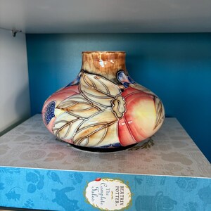 May include: A ceramic vase with a rounded form, decorated with a pattern of leaves and fruit in peach, pink, and brown hues. The vase is placed on a stack of books, with a blue background.