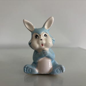 May include: A small ceramic figurine of a seated rabbit with a glossy finish. The rabbit is primarily light blue with a white chest, face, and inner ears. It has painted black eyes with long eyelashes, a small pink mouth, and pointed ears.