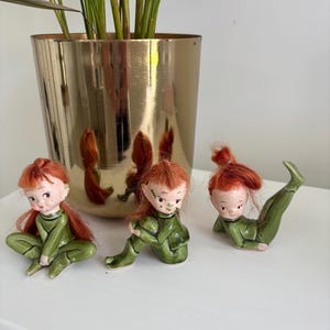 May include: Three ceramic elf figurines with red hair and green outfits are displayed on a white surface. The elves are in various poses, with one leg extended. A gold-coloured planter with a green plant is in the background.
