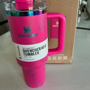 May include: A pink Stanley Quencher H2.0 tumbler with a handle and a clear lid. The tumbler has a pink lid and handle, with a silver band around the top. The tumbler has a white label with black text that reads "Quencher H2.0 Tumbler".