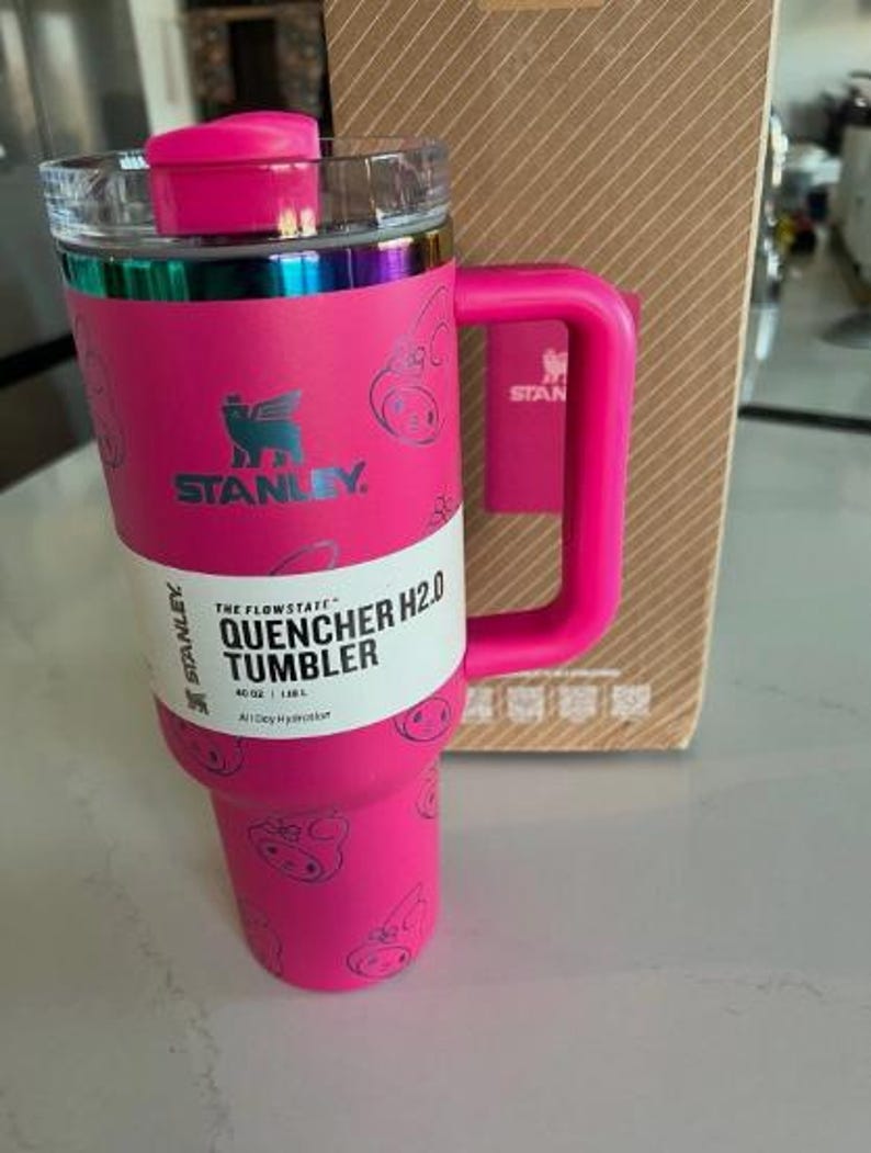 May include: A pink Stanley Quencher H2.0 tumbler with a handle and a clear lid. The tumbler has a pink lid and handle, with a silver band around the top. The tumbler has a white label with black text that reads "Quencher H2.0 Tumbler".