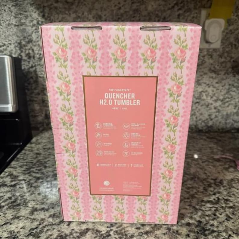 May include: A pink box with a floral pattern and the text "THE FLOW STATE QUENCHER H2.0 TUMBLER" in white. The box has a rectangular label with icons and text describing the product's features. The box is 11 inches tall.