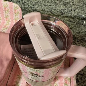 May include: A pink Stanley travel mug with a floral design. The mug has a handle and a white flip-top lid. The mug is made of a clear material and has a pink interior. The floral pattern includes pink roses and green leaves.