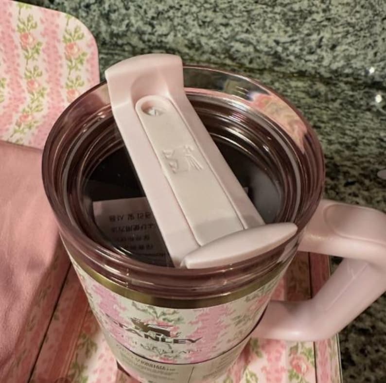 May include: A pink Stanley travel mug with a floral design. The mug has a handle and a white flip-top lid. The mug is made of a clear material and has a pink interior. The floral pattern includes pink roses and green leaves.