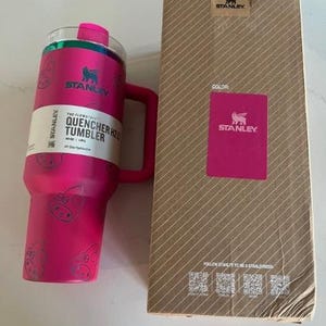 May include: A pink Stanley Quencher H2.0 tumbler with a handle and a matching pink box. The tumbler has a clear lid and a white label with black text. The box is brown with a pink Stanley logo.