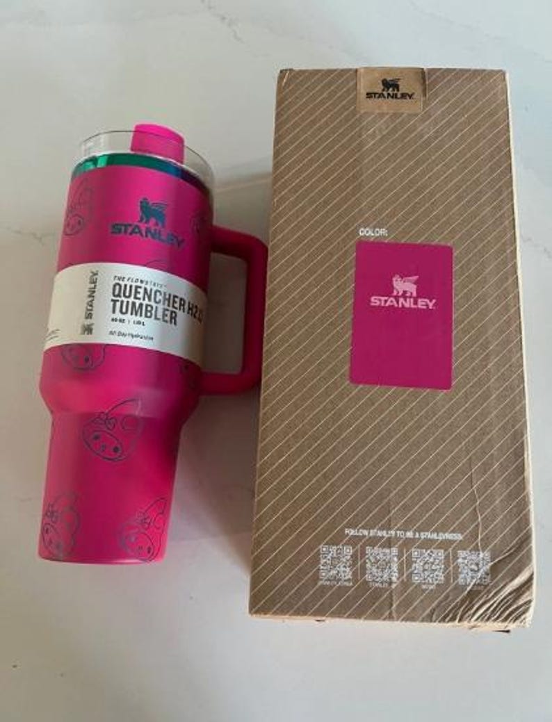May include: A pink Stanley Quencher H2.0 tumbler with a handle and a matching pink box. The tumbler has a clear lid and a white label with black text. The box is brown with a pink Stanley logo.
