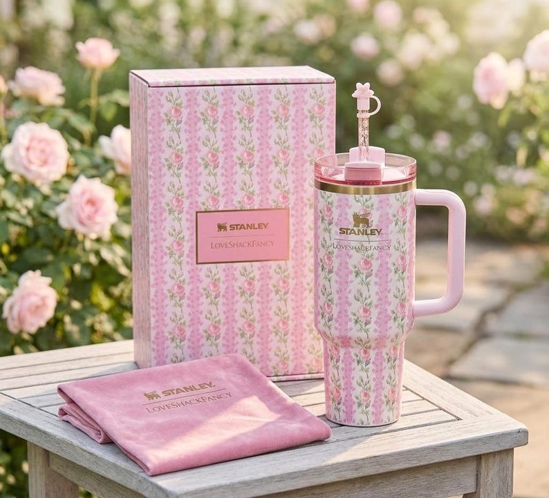May include: A pink floral-patterned Stanley tumbler with a handle, straw, and matching box and cloth. The tumbler and box feature a repeating pattern of pink roses and green leaves. The cloth is folded and has the Stanley and Loveshackfancy logos.