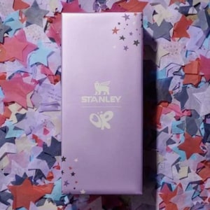 Puede incluir: Una caja rectangular de color lavanda con el logotipo "STANLEY" y un dise&ntilde;o de mariposa. La caja est&aacute; decorada con gr&aacute;ficos de estrellas y descansa sobre un fondo colorido de confeti en forma de estrella.