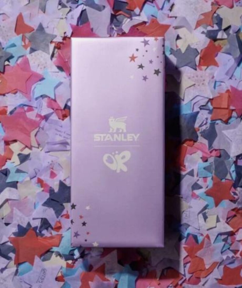 Puede incluir: Una caja rectangular de color lavanda con el logotipo "STANLEY" y un dise&ntilde;o de mariposa. La caja est&aacute; decorada con gr&aacute;ficos de estrellas y descansa sobre un fondo colorido de confeti en forma de estrella.