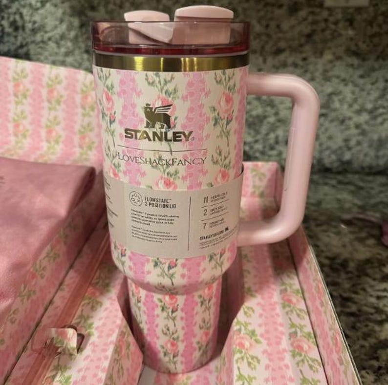 May include: A pink and white floral patterned Stanley Loveshackfancy tumbler with a pink handle and lid. The tumbler has a gold rim and the Stanley logo. The tumbler is in a matching floral patterned box.
