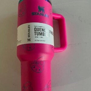 May include: A bright pink Stanley Quencher H2.0 Flowstate tumbler with a handle. The tumbler has a clear lid and is decorated with a repeating pattern of cartoon characters. The tumbler has a white label with the text "QUENCHER H2.0 TUMBLER" and "40 OZ | 1.18 L".