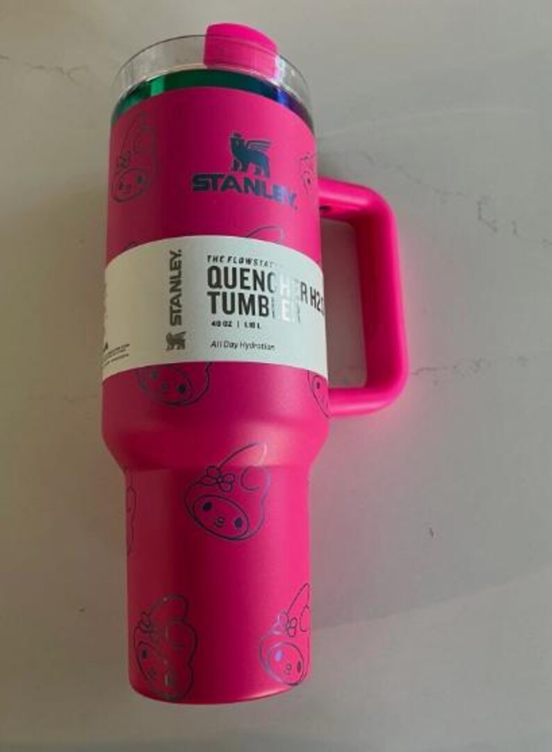 May include: A bright pink Stanley Quencher H2.0 Flowstate tumbler with a handle. The tumbler has a clear lid and is decorated with a repeating pattern of cartoon characters. The tumbler has a white label with the text "QUENCHER H2.0 TUMBLER" and "40 OZ | 1.18 L".