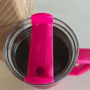May include: A clear glass tumbler filled with a dark liquid, possibly coffee or tea. The tumbler has a bright pink lid and handle. The lid features a small, embossed design and a circular button. The overall aesthetic is modern and functional.