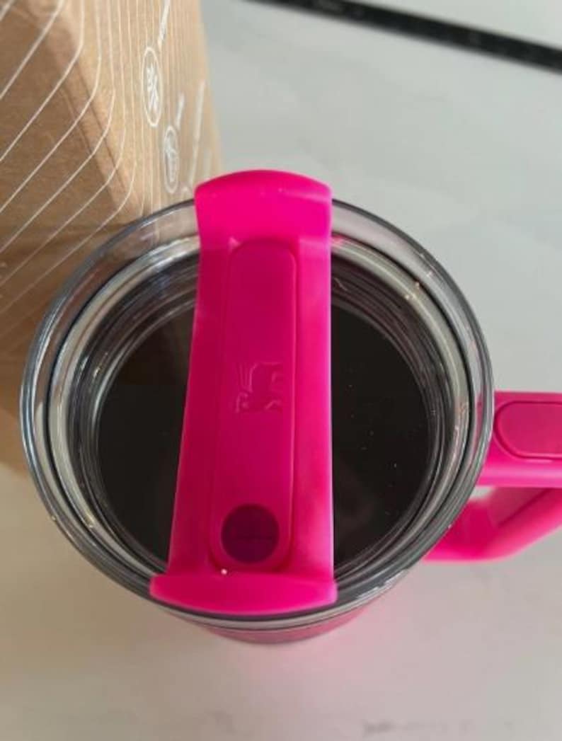 May include: A clear glass tumbler filled with a dark liquid, possibly coffee or tea. The tumbler has a bright pink lid and handle. The lid features a small, embossed design and a circular button. The overall aesthetic is modern and functional.