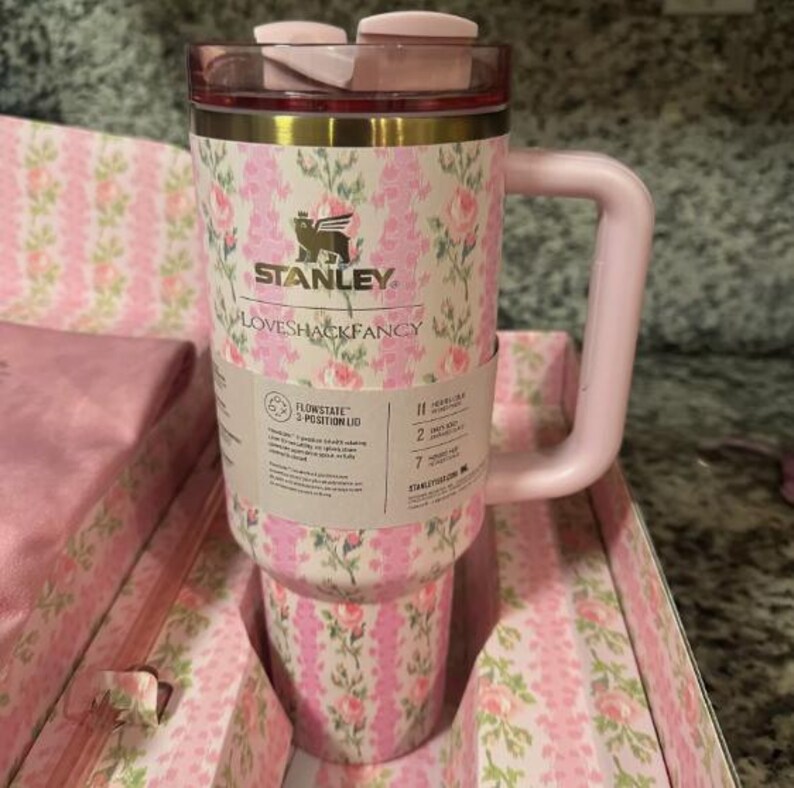 May include: A pink and white floral patterned Stanley Loveshackfancy tumbler with a pink handle and lid. The tumbler has a gold rim and the Stanley logo. The tumbler is in a matching floral patterned box.
