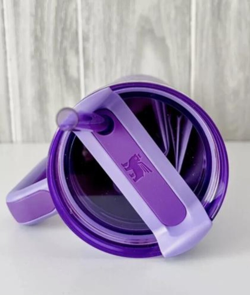 Puede incluir: Vaso morado con tapa y pajita a juego. La tapa presenta un asa morada clara y una banda morada oscura con un logotipo de caballo alado. Dise&ntilde;ado para bebidas fr&iacute;as.