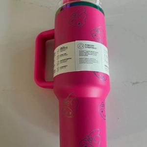 May include: A bright pink insulated tumbler with a handle and a clear lid. The tumbler is decorated with a repeating silver graphic of a cartoon pig with a bow. A white label with text is affixed to the side.
