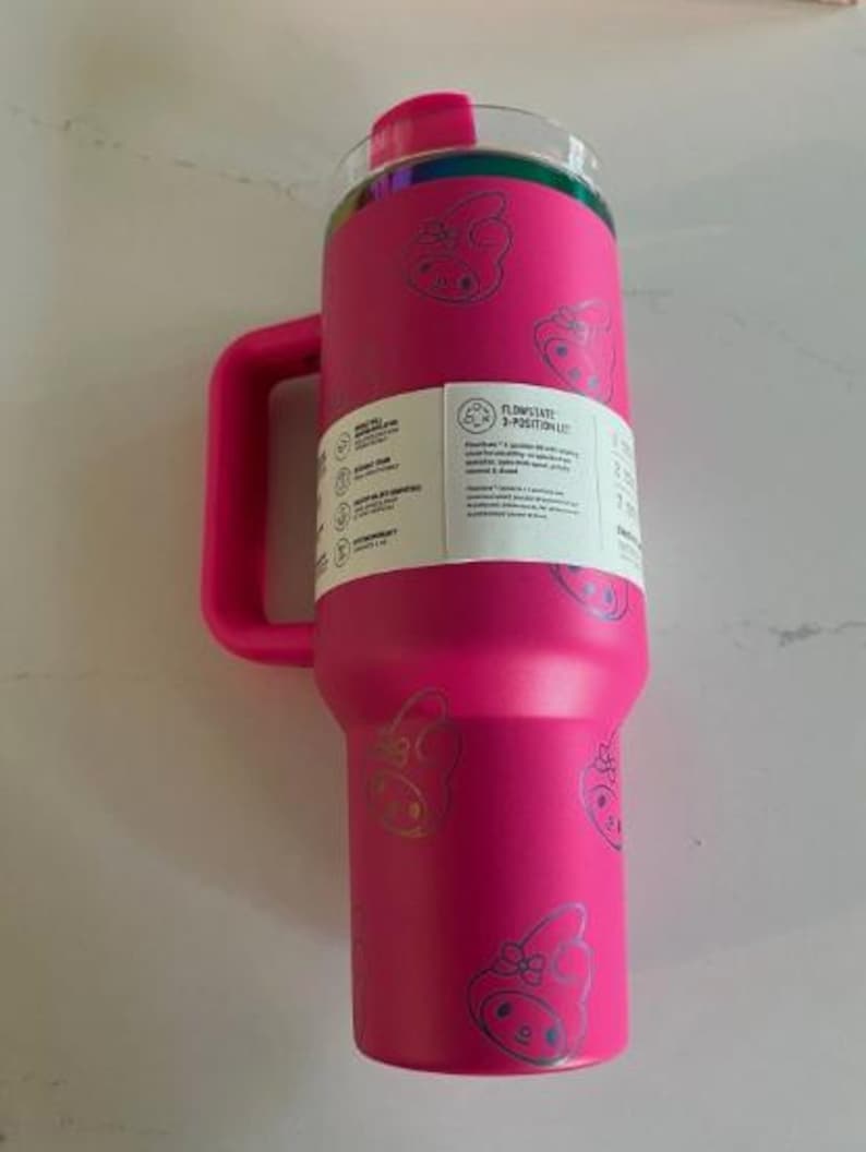 May include: A bright pink insulated tumbler with a handle and a clear lid. The tumbler is decorated with a repeating silver graphic of a cartoon pig with a bow. A white label with text is affixed to the side.