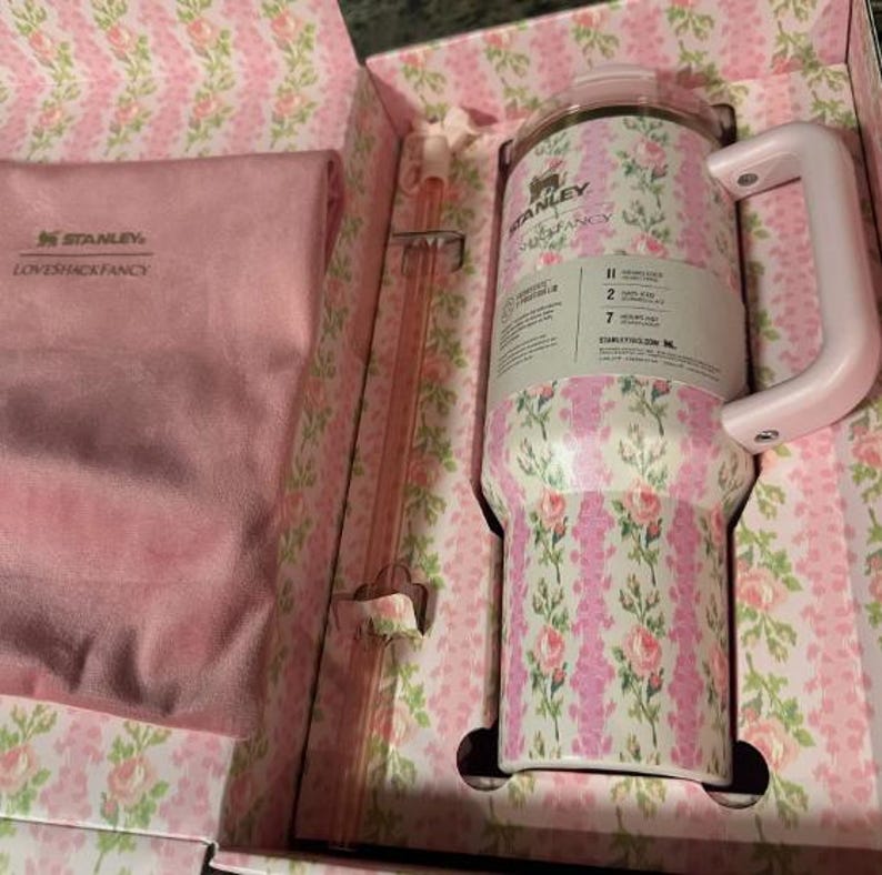 May include: A floral-patterned Stanley tumbler with a pink handle and lid, presented in a matching box. The tumbler features a design of pink roses and green leaves on a white background. A pink straw is also included.